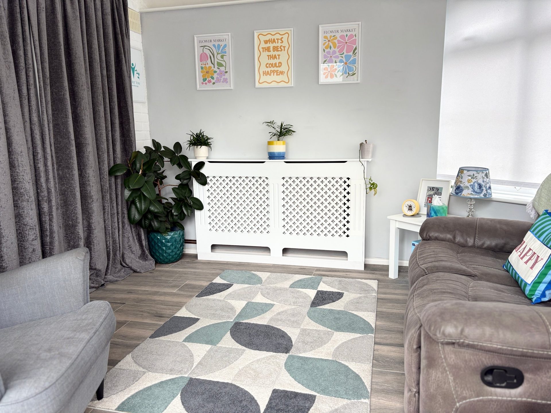 Bright, calm hypnotherapy room in Braintree with a grey armchair, a reclining sofa, plants and flower prints on the wall.