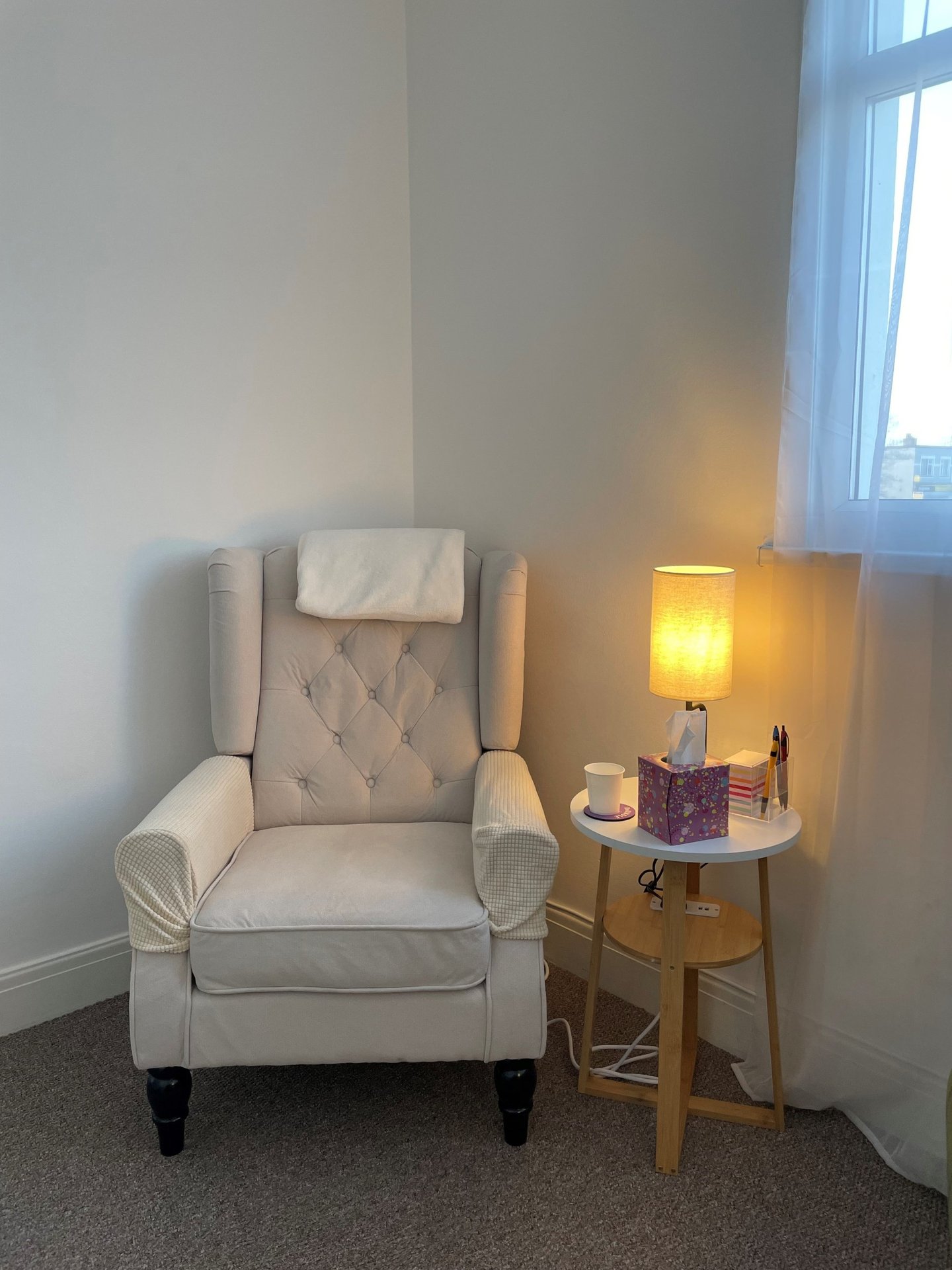 Cream wingback client chair beside a warm table lamp and tissues in Keeley Smith’s hypnotherapy room in Southend-on-Sea.