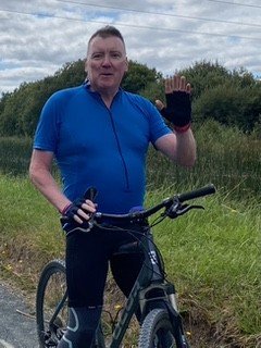 Peter Ely, hypnotherapist in Islington, waving from his bike on a country road.